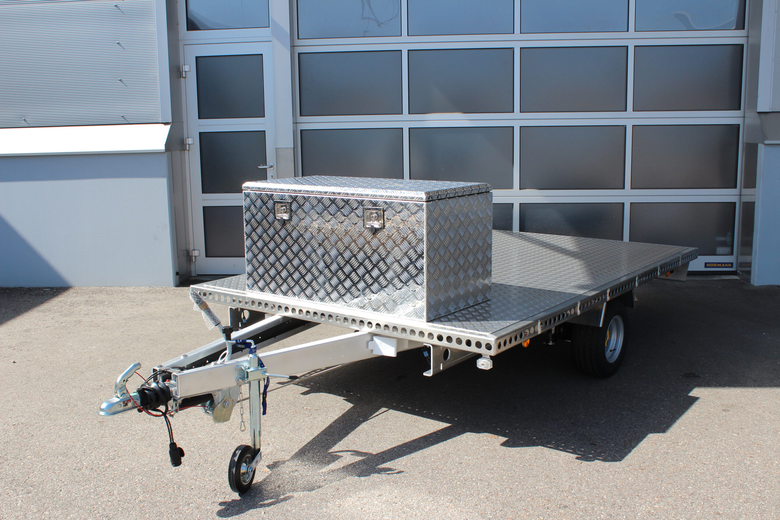 Große silberne Aluminium Riffelblech Staubox mit zwei Drehverschlüssen. Steht auf einem silbernen Aluminium Anhänger vor einem Tor