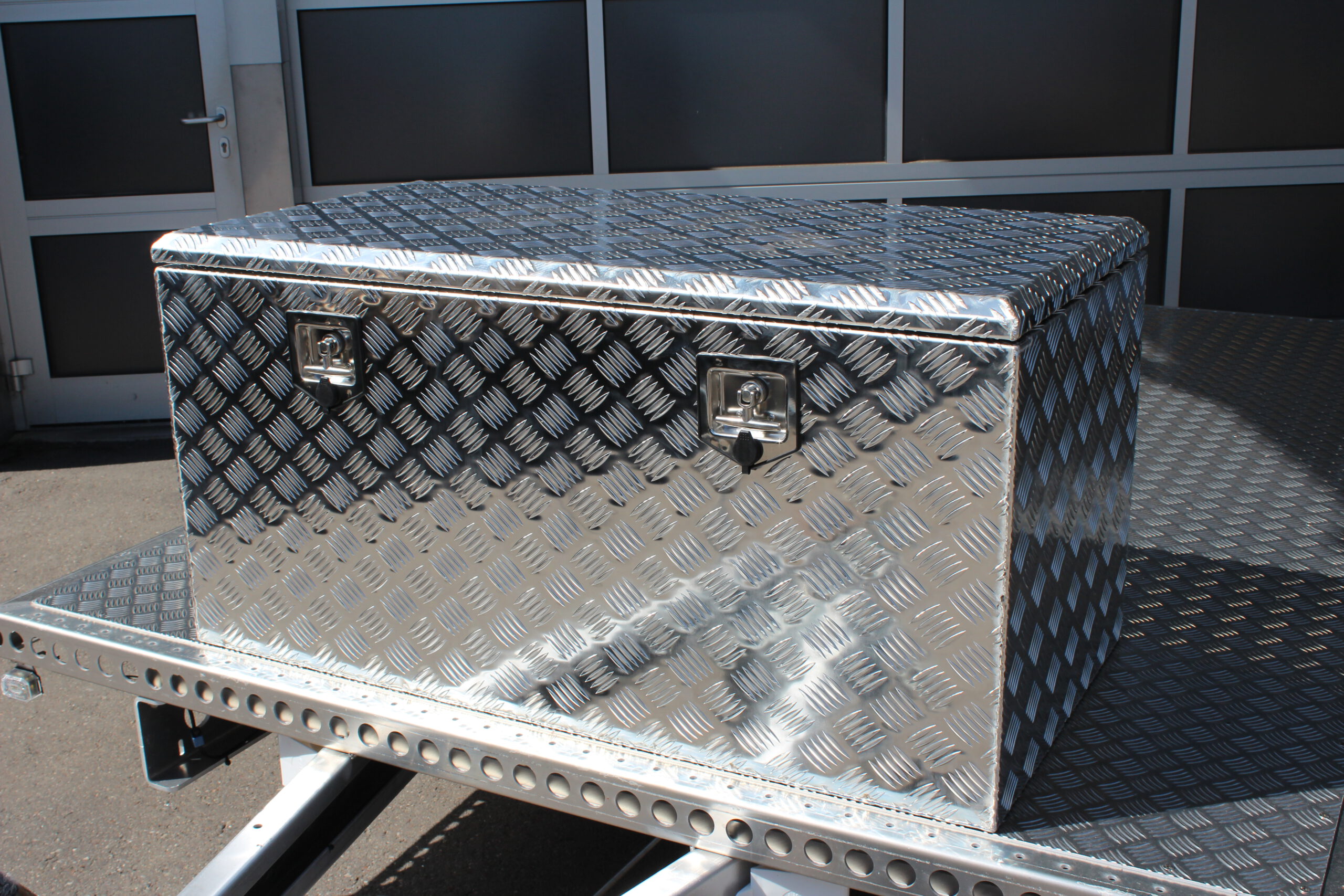 Große silberne Aluminium Riffelblech Staubox mit zwei Drehverschlüssen. Steht auf einem silbernen Aluminium Anhänger vor einem Tor