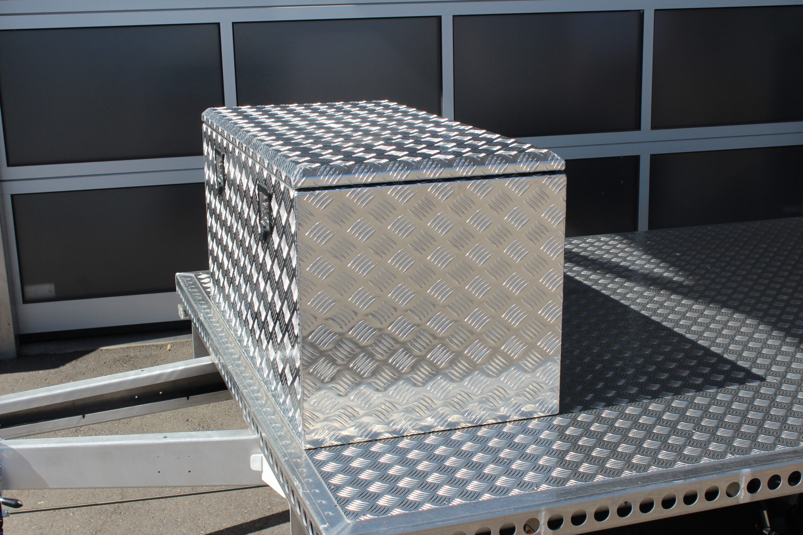 Große silberne Aluminium Riffelblech Staubox mit zwei Drehverschlüssen. Steht auf einem silbernen Aluminium Anhänger vor einem Tor