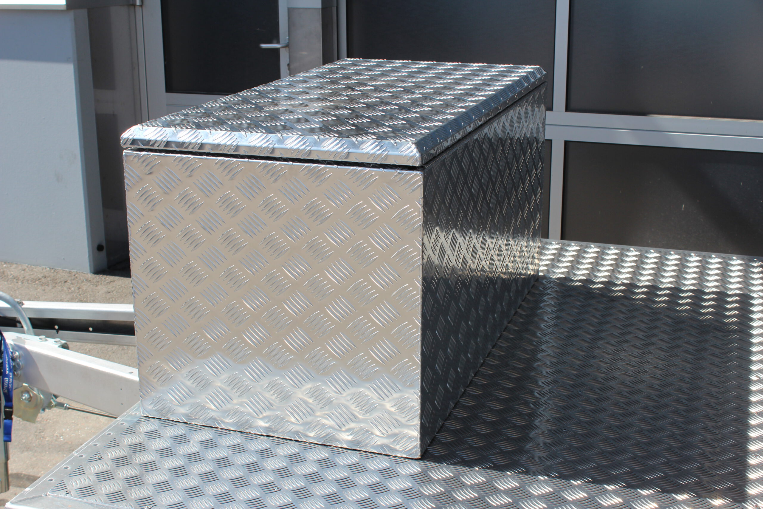 Große silberne Aluminium Riffelblech Staubox mit zwei Drehverschlüssen. Steht auf einem silbernen Aluminium Anhänger vor einem Tor