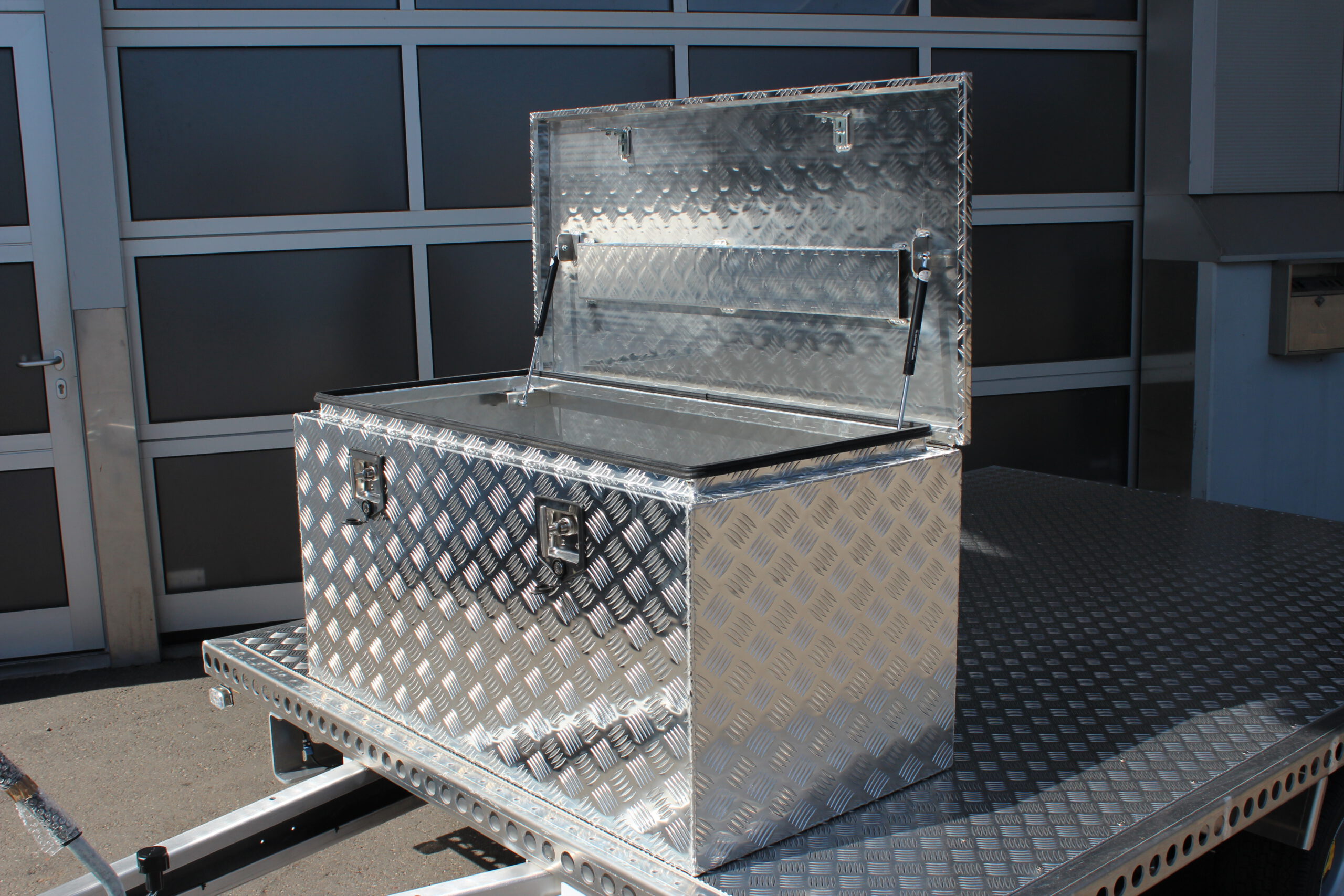 Große silberne Aluminium Riffelblech Staubox mit zwei Drehverschlüssen. Steht auf einem silbernen Aluminium Anhänger vor einem Tor