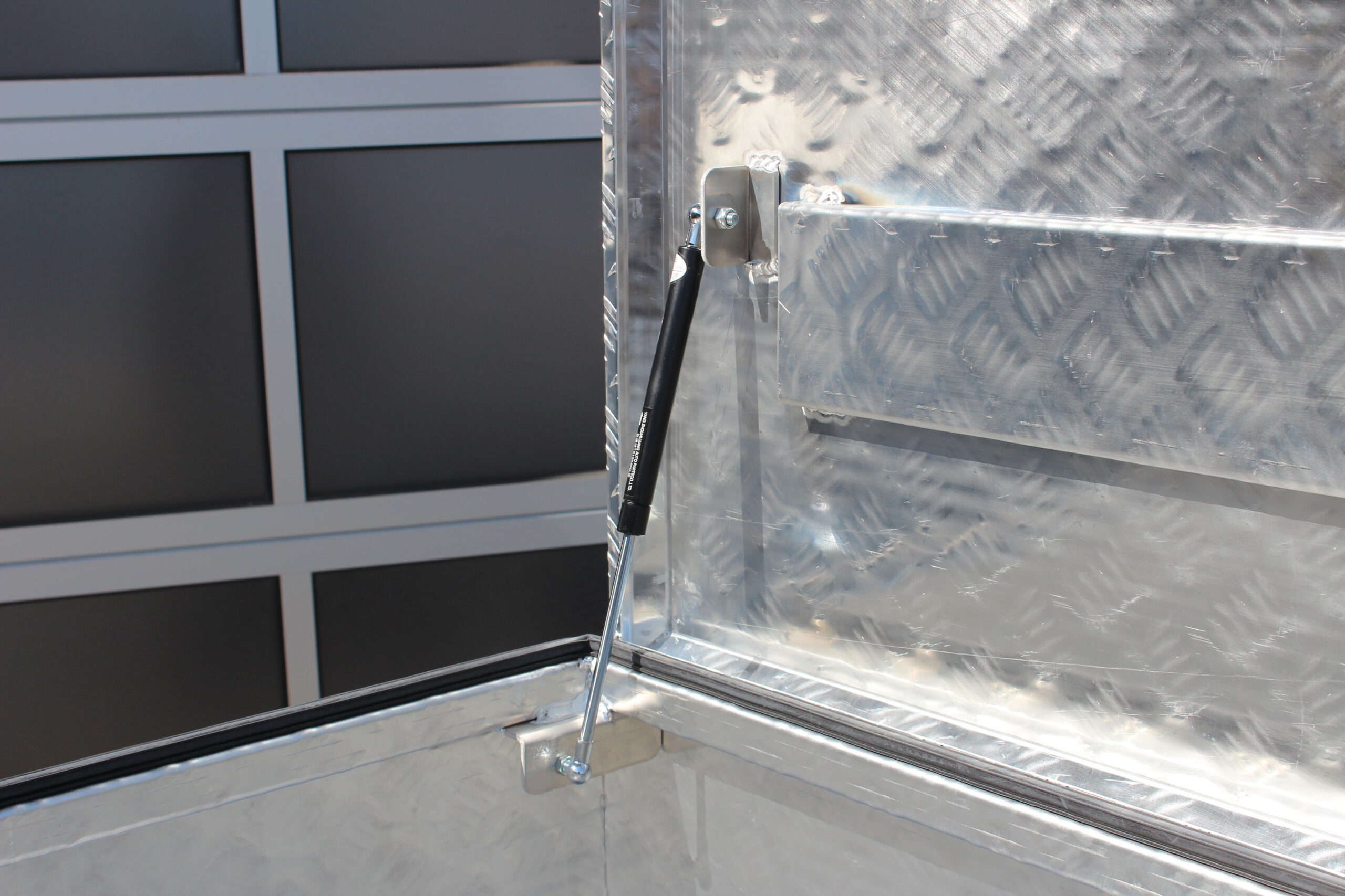 Große silberne Aluminium Riffelblech Staubox mit zwei Drehverschlüssen. Steht auf einem silbernen Aluminium Anhänger vor einem Tor